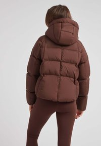Brązowa kurtka puchowa z kapturem, charakteryzująca się kwadratowym pikowaniem, grubym ociepleniem i krótkim krojem, zestawiona z dopasowanymi brązowymi legginsami.