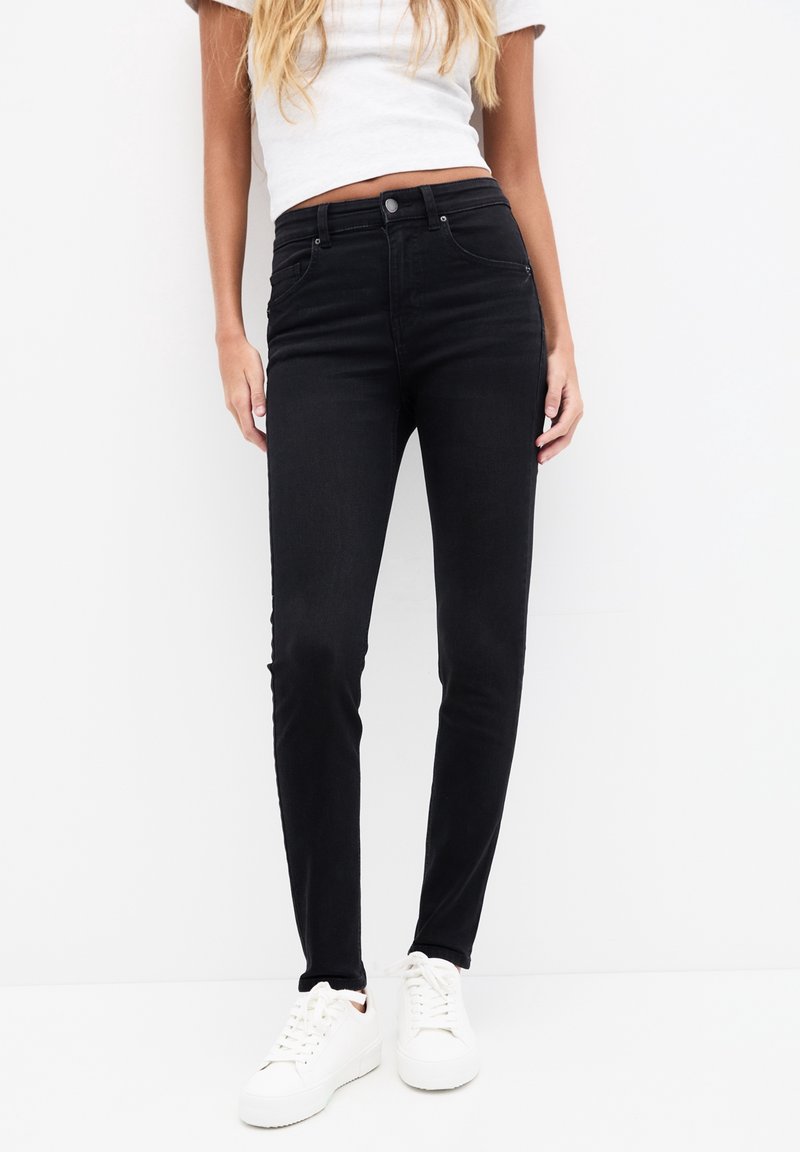 Jean skinny noirs à taille haute, en denim. Doté de poches classiques et de passants de ceinture, porté avec des baskets blanches.