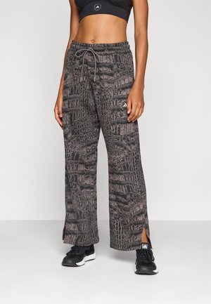 Femme portant un pantalon de survêtement ample avec imprimé crocodile noir et gris, taille élastique, et des chaussures de sport noires avec semelles blanches.