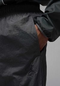 Czarne szorty przeciwwiatrowe z błyszczącej, teksturowanej tkaniny, elastycznym paskiem w talii, boczną kieszenią oraz czarnym logo Jumpman na lewym udzie.