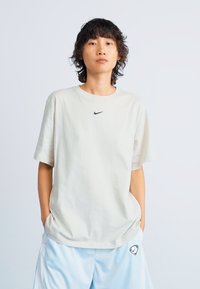 Šviesiai béžinė Nike marškinėliai su apvalia apykakle, trumpomis rankovėmis ir mažuoju juodu logotipu ant krūtinės. Dėvimi su šviesiai mėlynomis kelnėmis.
