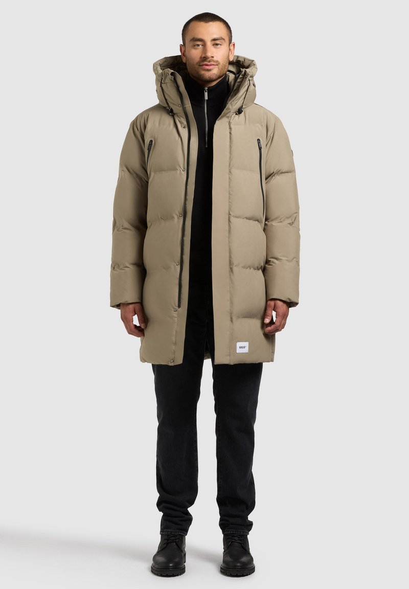 Abrigo de plumas beige con capucha, con un diseño acolchado y bolsillos con cremallera en negro. Lleva puesto sobre una camiseta negra y combinado con pantalones oscuros.