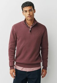 Bordeauxfarbener Pullover mit einem Reißverschluss-Halstecker, gerippten Bündchen und Saum. Über einem gestreiften, mit Kragen versehenen Hemd getragen. Glatte Textur, lässiges Design.