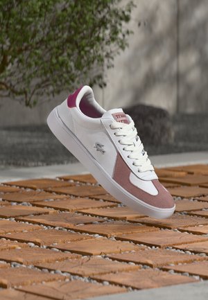 Fehér és rózsaszín cipőfűzős sportcipő lebeg a texturált fa burkolat felett kültéren, háttérben zöld növényzettel és betonfallal.