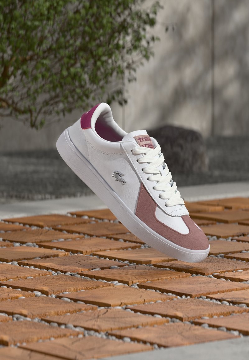 White and pink sneaker with laces floating above textured wooden tiles outdoors, with greenery and concrete wall in the background.