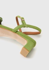 Sandalo verde in suede con tacco quadrato. Presenta una sottile cinturino alla caviglia con fibbia metallica e un'intersuola testurizzata. Accenti in pelle marrone visibili.