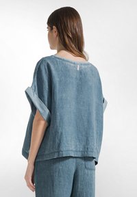 Donna con top in denim azzurro chiaro a maniche larghe arrotolate e pantaloni abbinati, vista da dietro su sfondo bianco uniforme.