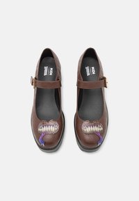Brązowe buty z ekoskóry w stylu Mary Jane, z charakterystycznym graficznym motywem potwora w kolorze purpurowym i białym na czubku oraz złotym akcentem w postaci klamry.