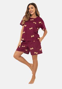 Uśmiechnięta kobieta stoi boso, ubrana w dopasowany bordowy t-shirt z krótkim rękawem i szorty w żółte wzory kokardek, z ręką w kieszeni.