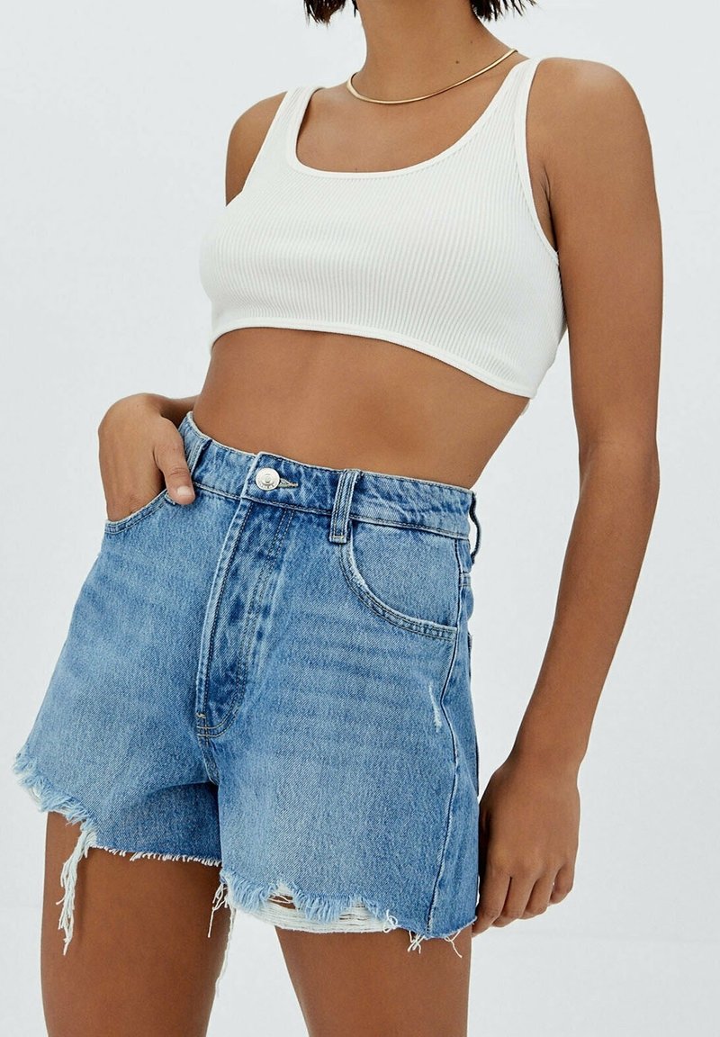 Femme portant un crop top blanc sans manches et un short en denim bleu taille haute avec des ourlets effilochés sur un fond clair.