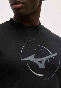 Κοντινό πλάνο ενός ατόμου που φοράει μαύρο πουκάμισο με μεγάλο, αφηρημένο ασημένιο γεωμετρικό σχέδιο στο στήθος, με απλό φόντο.