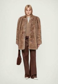 Imitatiebontjas in lichtbruin met een losse pasvorm. Gecombineerd met een beige trui en bruine broek, aangevuld met een kleine bruine tas.