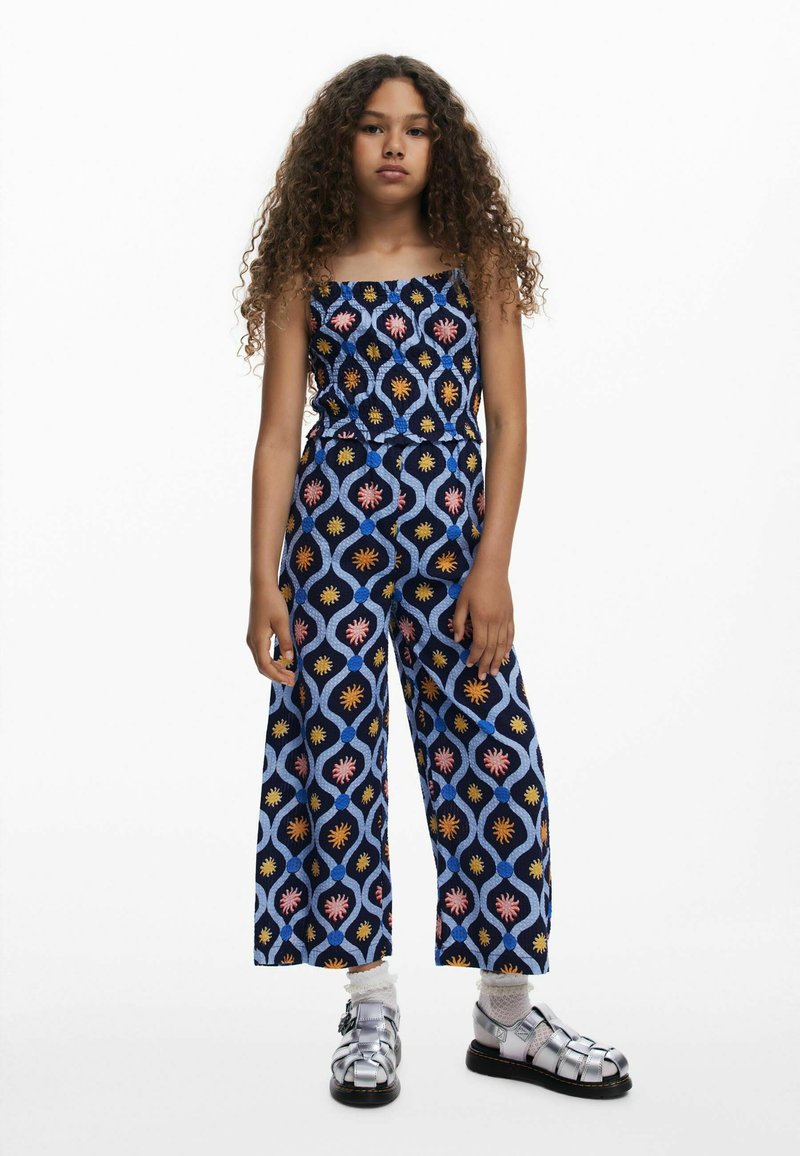 Blue patterned jumpsuit with floral designs in pink, yellow, and white; wide legs and strapless top. Model wears silver sandals with white socks.