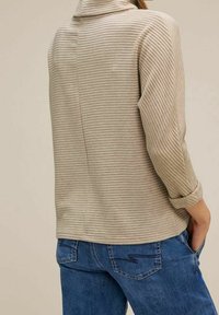 Beżowy, prążkowany top z długim rękawem o wysokim kołnierzu i podwiniętych rękawach, w połączeniu z niebieskimi dżinsami. Tył ma szew biegnący wzdłuż środka.