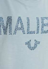 Světle modré tričko s třpytivými modrými kamínky, které tvoří slovo "MALIBU" a dekorativním akcentem pod ním, připomínajícím dvojitou šipku.