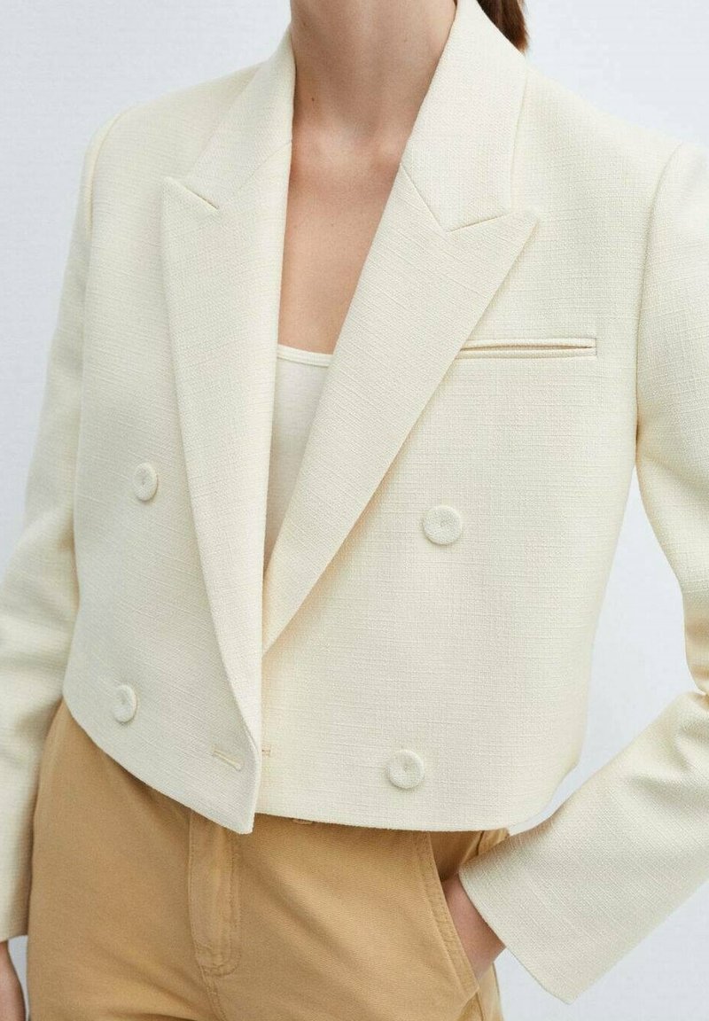 Femme portant un blazer court croisé crème en tissu texturé sur un pantalon beige, main dans la poche, sur un fond uni.