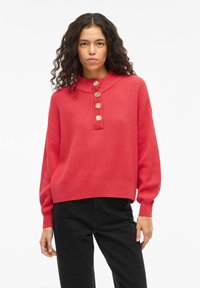 Punane soonikkoega kampsun kõrge kraega, viie suure nupuga ja pikkade varrukatega. Kandmiseks mustade pükstega, mis on lõdva istumise ja lühendatud pikkusega.