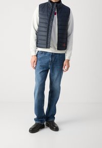 Granatowa pikowana kamizelka puchowa nałożona na jasnoszary sweter, w połączeniu z niebieskimi dżinsami i czarnymi botkami. Posiada zamek błyskawiczny oraz akcent z logo.