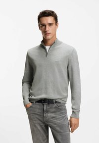 Szary sweter z suwakiem przy kołnierzu, wykonany z miękkiego materiału. Noszony na białej koszulce, zestawiony z ciemnoszarymi jeansami. Subtelne logo na piersi.