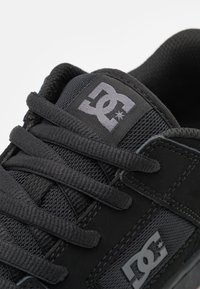 Fekete sneaker, textúrált bőrből és hálós anyagból készült felsőrésszel, lapos fekete fűzőkkel és a szónokoldalán kiemelkedő szürke DC logóval.