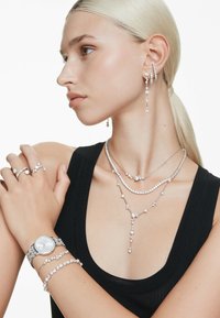 Silberschmuck umfasst geschichtete Halsketten, Ohrringe, eine Uhr und Ringe, die mit funkelnden Steinen verziert sind, an einem Modell vor einem weißen Hintergrund.