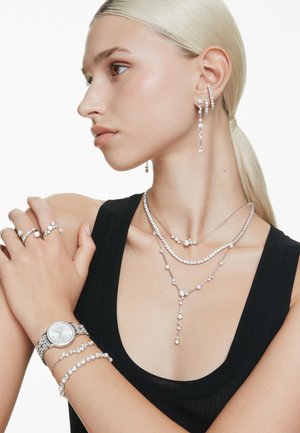 Silver jewelry includes layered necklaces, drop earrings, a watch, and rings embellished with sparkling stones on a model against a white background.