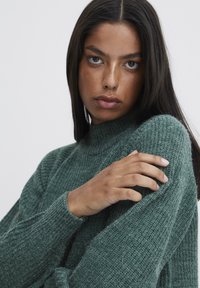 Strickpullover in tiefgrün mit einem hohen Kragen und strukturiertem Stoff sowie gerippten Bündchen. Hand sanft auf der Schulter platziert.
