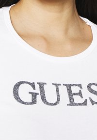 Camiseta blanca con cuello redondo y texto en purpurina plateada que dice "GUESS" en el pecho. Material de algodón suave, textura lisa.