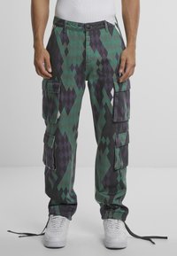 Pantalones cargo con un patrón de diamantes en verde y negro, que cuentan con múltiples bolsillos, un corte relajado y bajos con cordón. Se llevan con zapatillas blancas.