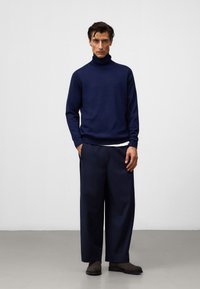 Navyfarbener Rollkragenpullover mit gerippten Bündchen und Saum, kombiniert mit weit geschnittenen navyfarbene Hosen und dunklen Wildlederschuhen. Schlichte, cleane Ästhetik.