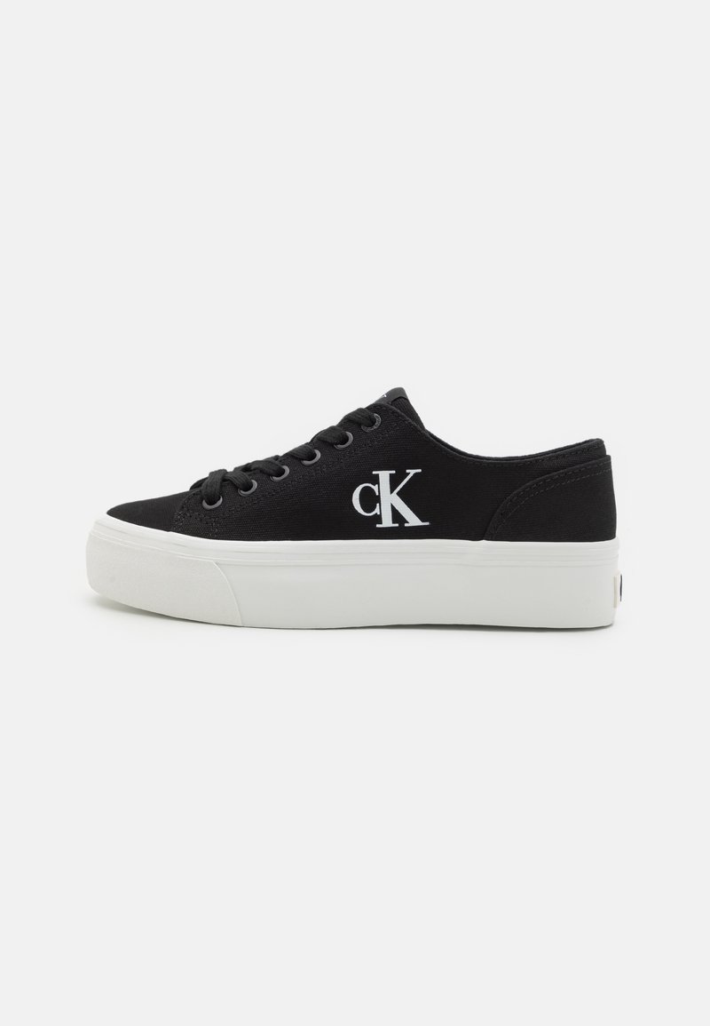 Schwarze Leinensneaker mit runder Spitze, weißer Gummisohle und kontrastierendem weißen Logo "cK" an der Außenseite. Leichtes Design.