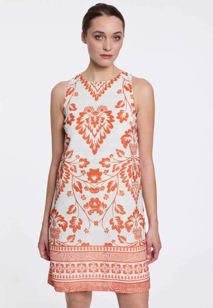 Femme portant une robe blanche sans manches avec des motifs floraux et géométriques orange, debout devant un fond clair uni.