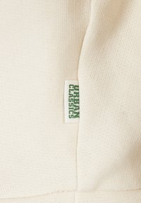 Detailný záber na pletenú látku v krémovej farbe s malou bielou etikettou s textom "URBAN CLASSICS" v zelenej farbe.