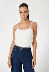 White, fuzzy cropped tank top with spaghetti straps and soft texture, paired with high-waisted dark denim pants, featuring a button closure.