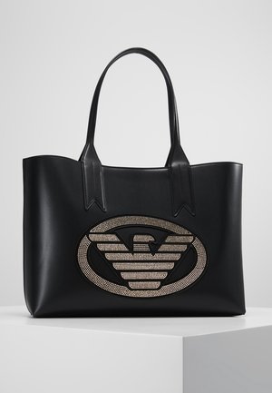 Bolso tote de cuero negro con dos asas y un gran emblema ovalado que presenta un logo de águila con tachuelas en el frente.