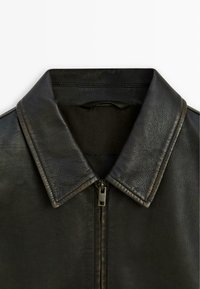 Black leather jacket featuring a wide collar and zip closure, with textured finish and visible stitching detail. Design emphasizes simplicity.