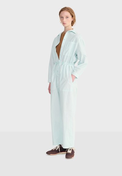 Jeune femme portant une combinaison à manches longues bleu clair avec une taille cintrée, un chemisier marron en dessous et des baskets marron foncé, debout de profil.