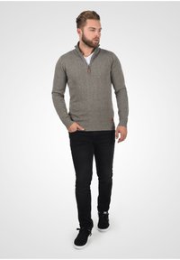 Maglione grigio a maglia con colletto a zip a un quarto, polsini e orlo a costine, e una toppa marrone con il marchio in vita, abbinato a jeans neri e sneakers.