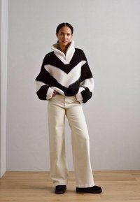 Suéter de punto en blanco y negro, con un patrón en forma de chevrón, cuello alto, corte oversize y puños acanalados. Combinado con pantalones de pierna ancha color crema.