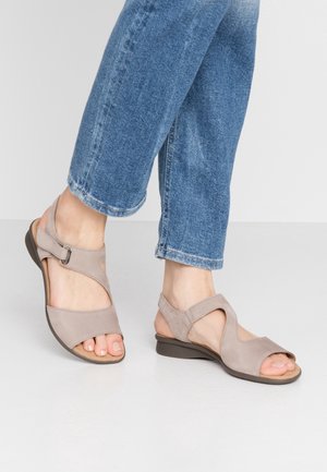 Person iført beige åbne sandaler med stropper og blå jeans, stående på en lystfarvet gulv.