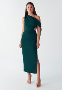 Abito turchese monospalla con dettaglio cut-out, vita arricciata, spacco laterale e tessuto liscio. Abbinato a sandali dorati con cinturini.