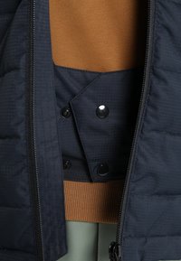 Veste matelassée bleu foncé partiellement dézippée sur un pull marron et un pantalon vert clair, montrant le détail du rabat intérieur boutonné.