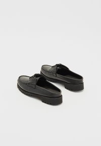 Chaussures noires en cuir sans lacets avec un design mocassin, présentant une couture sur le dessus, une semelle basse et une semelle extérieure en caoutchouc texturé.