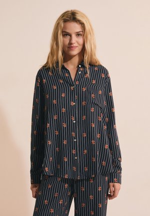 Blonde Frau trägt einen dunkelblauen gestreiften Pyjama mit kleinen braunen Bärengesichtern und steht vor einem hellbeigen Hintergrund.