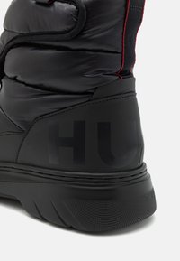Bota aislante negra con acabado brillante, que presenta una suela texturizada, acentos de costura roja y un prominente logo "HU" en el talón.