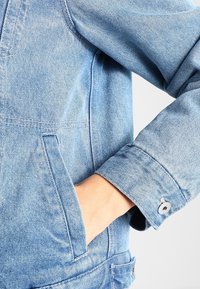 Giacca di jeans azzurro chiaro con una tasca sul lato sinistro e dettagli nei polsini. Il tessuto presenta una texture sbiadita e cuciture prominenti.