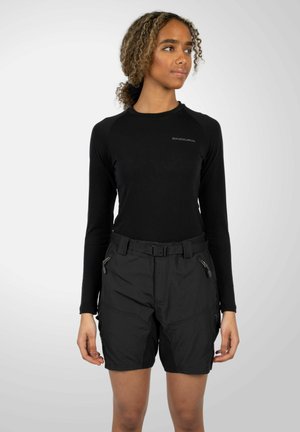 Femme portant un haut noir moulant à manches longues et un short noir ceinturé avec des poches zippées, regardant légèrement sur le côté sur un fond clair uni.