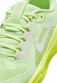 Chaussure de sport en maille verte avec une tige texturée, accents blancs, un col rembourré et une semelle jaune robuste. Lacets et détails de ventilation visibles.