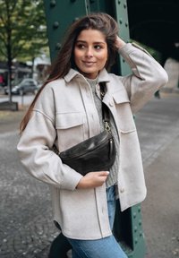 Woman in beige jacket and gray sweater holding a black crossbody bag, standing by a green metal pillar on a city street.