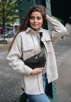 Woman in beige jacket and gray sweater holding a black crossbody bag, standing by a green metal pillar on a city street.